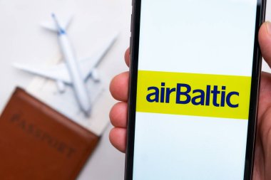 AirBaltic Havayolları uygulaması akıllı telefon ekranının elinde. Beyaz oyuncak bir uçak ve pasaport, hafif bir yüzeyi olan bir masada yatıyor. Kasım 2021, San Francisco, ABD.