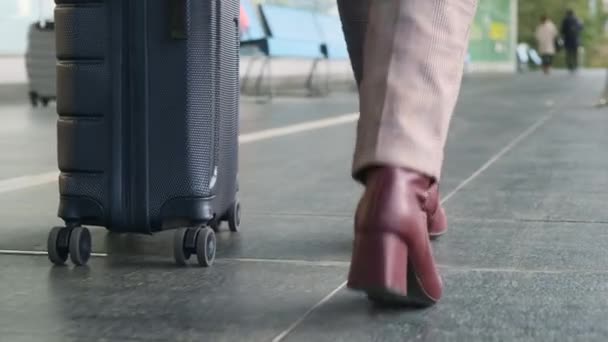 Женщина в светлых брюках, в бордовых туфлях на каблуках, с серым чемоданом на колесах медленно выходит на улицу вдоль терминала аэропорта и пустыми скамейками, с видом на ноги снизу. Концепция путешествия, деловая поездка — стоковое видео