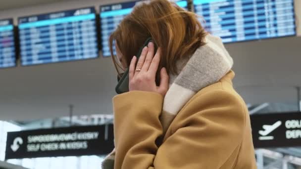 Jonge bruinharige vrouw met zwart gezichtsmasker praat op mobiele telefoon tegen de achtergrond van aankomst en vertrekbord in het luchthavengebouw, globaal mobiel communicatieconcept. Zijaanzicht. — Stockvideo