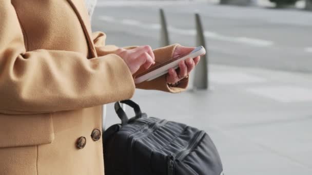 Εγκύκλιος flyby της κάμερας γύρω από γυναίκα χωρίς πρόσωπο αναζήτηση πληροφοριών στο smartphone, σε εξωτερικούς χώρους στο χώρο στάθμευσης του αεροδρομίου, φορώντας μοντέρνα μπεζ παλτό κασμίρ. Έννοια της εφαρμογής για κινητά ταξί — Αρχείο Βίντεο