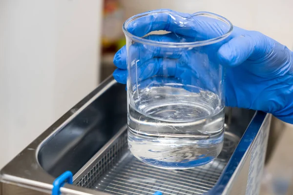 Επιστήμονας σε μπλε καουτσούκ βάζει ένα ποτήρι για διάλυση ή καθαρισμό σε ένα λουτρό υπερήχων. Κλινική, τοξικολογική και ιατροδικαστική ανάλυση — Φωτογραφία Αρχείου