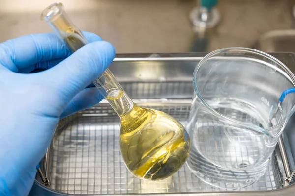 Close up assistente de laboratório mão em luva azul, segurando frasco de vidro redondo líquido amarelo, recipiente de caixa de metal, copo de medição transparente copo, derramando cuspir. — Fotografia de Stock