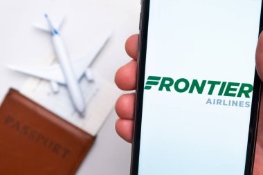 Frontier Havayolları uygulaması akıllı telefon ekranında gösteriliyor. Arka planda bulanık bir uçak, pasaport ve biniş kartı var. Kasım 2021, San Francisco, ABD.