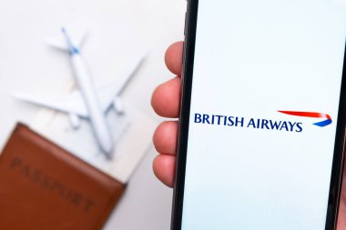 Cep telefonu ekranında İngiliz havayolları uygulama logosu var. Arka planda bulanık bir uçak görüntüsü, pasaport ve biniş kartı. Kasım 2021, San Francisco, ABD.