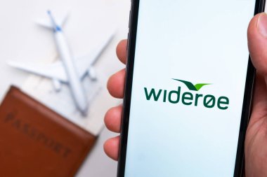 Wideroe Havayolları uygulaması cep telefonu ekranında arka planda uçak, uçuş kartı ve pasaport olan adamlar var. Havayolları mobil uygulaması kavramı. Kasım 2021, San Francisco, ABD.