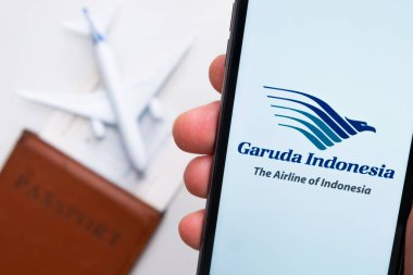 Garuda Endonezya Havayolları uygulaması veya logosu arka planda pasaport, uçuş kartı ve uçak olan bir cep telefonunda görüntülendi.