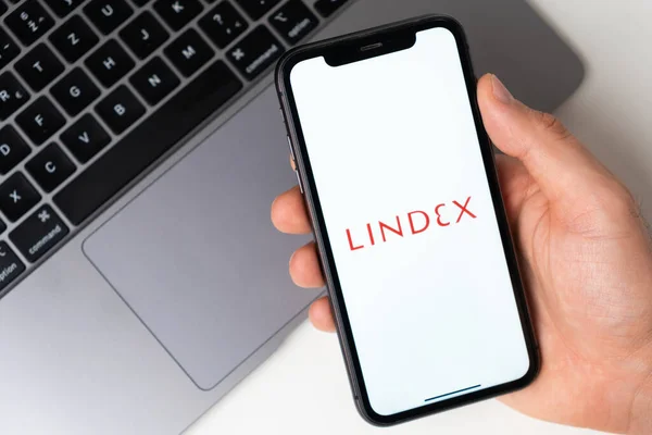 手机屏幕上的Lindex应用在男人手里，背景是笔记本电脑。2021年11月在美国旧金山通过手机进行网上购物 — 图库照片