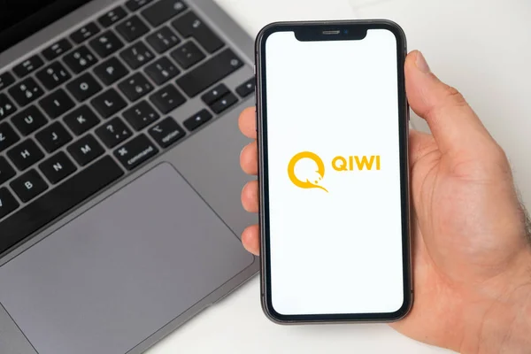 Plateforme financière Qiwi pour le transfert d'argent à l'aide d'un smartphone. Homme main tenant téléphone portable avec application sur l'écran et ordinateur portable sur l'arrière-plan. Novembre 2021, San Francisco, États-Unis — Photo