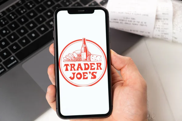 Trader Joes application on the screen of Apple iPhone in mans hand and laptop on the background. Online shopping concept November 2021, San Francisco, USA