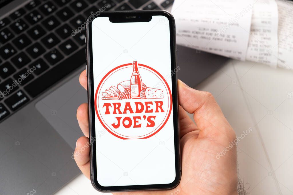 Trader Joes application on the screen of Apple iPhone in mans hand and laptop on the background. Online shopping concept. November 2021, San Francisco, USA