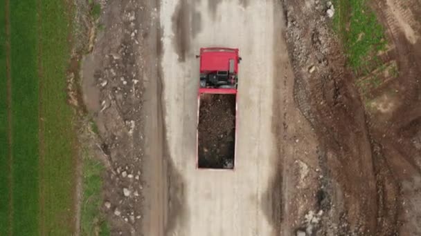 2つの緑のフィールドの間の未舗装の道路に沿ってフィールドから土壌を運ぶ赤いローリー — ストック動画