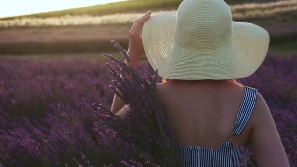 Cinematic footage of woman stands with her back, holds a bouquet of lavender and slowly touches her hat with her hand in lavender field at sunset — Stockvideo