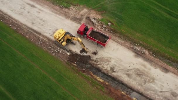 A yellow excavator CAT loads soil from the field into a red truck Tatra during excavation work and loaded truck driving away. Heavy earth moving equipment loading, lifting and transportation. October — Wideo stockowe