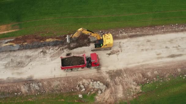 Una excavadora CAT amarilla carga el suelo del campo en un camión rojo durante el trabajo de excavación y un camión cargado alejándose. Carga, elevación y transporte de equipos de movimiento de tierra pesada, octubre de 2021 — Vídeos de Stock