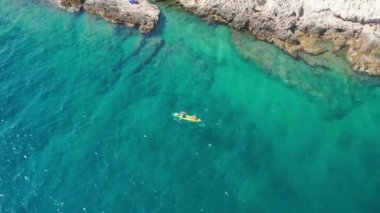 Drone footage of two people are sailing in a kayak along a rocky coast with clear sea water and a rocky bottom is visible at a depth