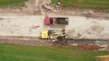Top view of a yellow excavator loads soil from the field into a red truck during excavation work. Heavy earth moving equipment loading, lifting and transportation