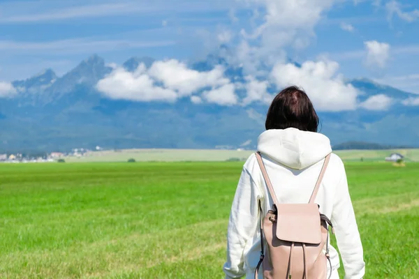 A woman tourist in a white hoodie and a backpack enjoys a beautiful view of the Tatra mountains while standing in a green field. Clouds float over mountain peaks, copy space. — стоковое фото
