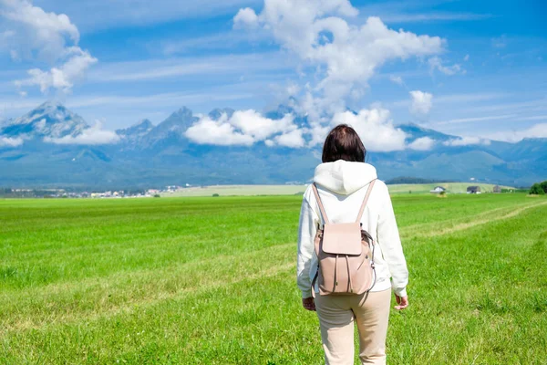 A woman tourist in a white hoodie and a backpack enjoys a beautiful view of the Tatra mountains while standing in a green field. Clouds float over mountain peaks, copy space. — стоковое фото