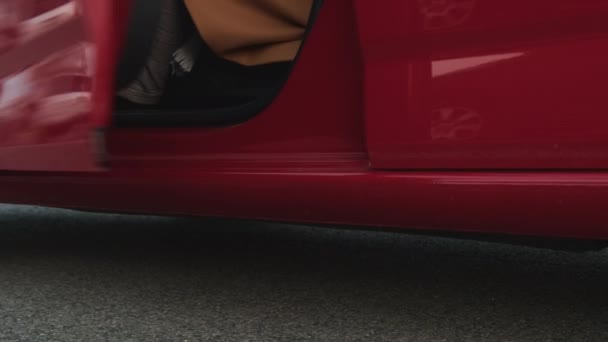 Una mujer de negocios en pantalones a cuadros, botas de color burdeos y un abrigo marrón abre la puerta de un coche rojo y sale — Vídeo de stock
