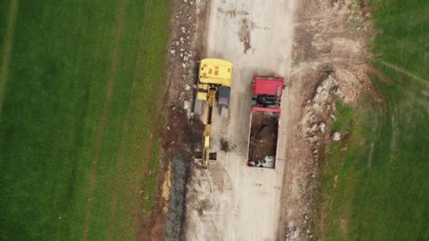 Πάνω όψη κίτρινο εκσκαφέας παίρνει γη από το πεδίο και το φορτώνει σε ένα κόκκινο φορτηγό — Αρχείο Βίντεο