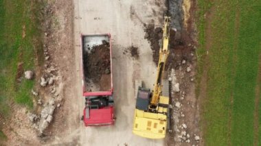 Üstteki sarı kazıcı tarladan arazi alıp kırmızı bir kamyona yüklüyor.