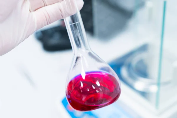 Frasco redondo con una muestra de sangre en manos de científicos en guantes de goma en un laboratorio moderno con una balanza analítica en el fondo — Foto de Stock