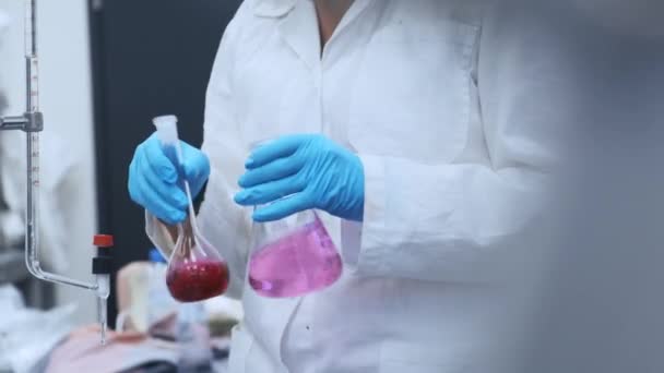 Cientista em luvas de borracha e jaleco misturando líquidos rosa nos frascos de vidro, realização do experimento. Laboratório moderno farmacêutico ou químico. — Vídeo de Stock