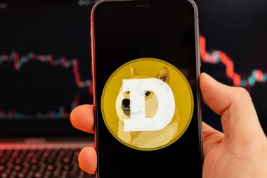 Kripto döviz piyasası analizinin Dogecoin DOGE uygulaması insan eliyle cep telefonu ekranında ve düşüş grafiklerinde arka planda veri alışverişi, Şubat 2022, San Francisco, ABD