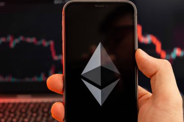 İnsan eliyle cep telefonu ekranındaki kripto döviz piyasası analizinin Ethereum ETH uygulaması ve arka plandaki eğilim grafikleri, Şubat 2022, San Francisco, ABD