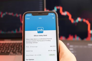 Silikon Vadisi Bankası borsada düşüş gösteriyor ve arka planda grafikler çizelgesi var. Şirket logosu olan bir cep telefonu taşıyan adam, Şubat 2022, San Francisco, ABD