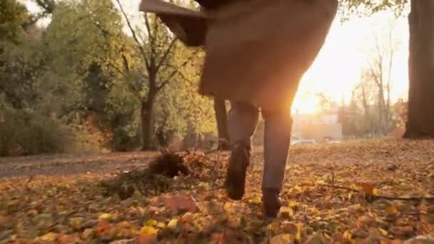 Жінка в пастельному одязі і коричневе пальто біжить на опале листя восени на заході сонця — стокове відео
