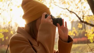 Kadın fotoğrafçı şık bir şekilde giyinmiş kameraya odaklanma ayarları yapıyor ve günbatımında sonbahar parkının fotoğrafını çekiyor.