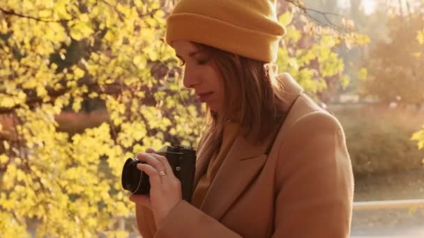 Mujer fotógrafa en sombrero naranja y abrigo ajusta el enfoque en la cámara para una imagen en el parque de otoño — Vídeo de stock