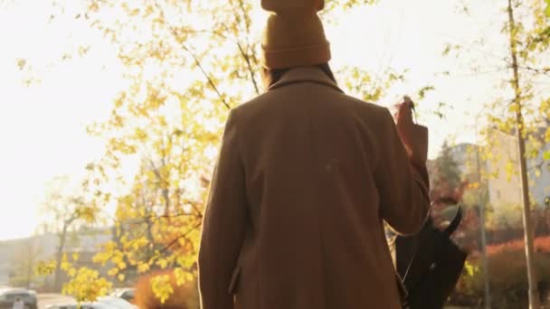 Stylově oblečená dívka si přehodí přes rameno batoh, zamává rukou a odchází do západu slunce — Stock video