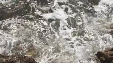 Köpüklü deniz dalgaları parlak güneş ışığında sahilde kayaların üzerinde süzülür.