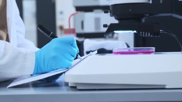 Cientista de perto em luvas de borracha fazendo anotações da experiência microbiológica. Placa de Petri com a amostra está sob microscópio. — Vídeo de Stock