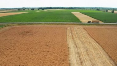 Tractor with hay and cars drive along road past green and yellow fields with harvest. Automobiles and trucks drive on asphalt in countryside aerial view