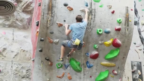 The climber trains on the artificial rock wall with insurance in bouldering gym — Stock Video