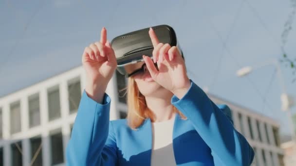 A happy woman in the suit is standing outside and gestures in augmented reality in 3D VR goggles — Stok Video