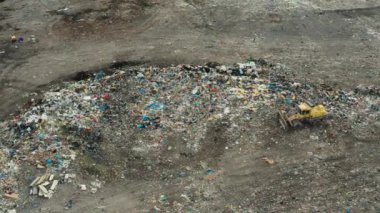 Buldozer, kırsal alanda çöp yığını olan büyük bir çöp yığınının yanında duruyor. Kuşlar atık ve plastik hava manzaralı evlerin üzerinde uçar.