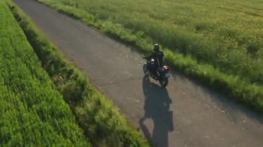 Motosikletliler tarlalar arasındaki küçük asfalt yolda hızlı sürerler. Motosikletli, büyük askı manzaralı kırsal eklentiden geçerken bisiklet sürüyor.