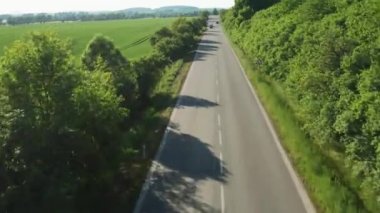 Cars and vehicles drive on road between green planted field and dense forest. Trees cast shadows on asphalt countryside road aerial view