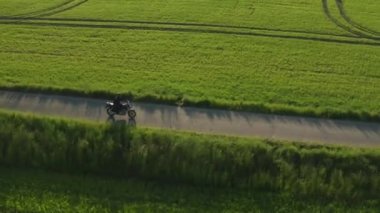 Motorcyclist rides on countryside road past green fields and trees at sunset. Small green bushes cast shadows on empty road aerial view