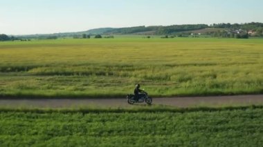 Motorcyclist rides on asphalt road past green field at sunset light. Fine weather at bright sunshine lighting fields against hills aerial view