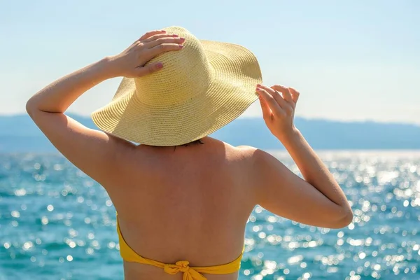 Elegant lady in yellow bikini admires sparkling sea at resort at sunlight. Young woman holds straw hat with hands on windy summer day close back view