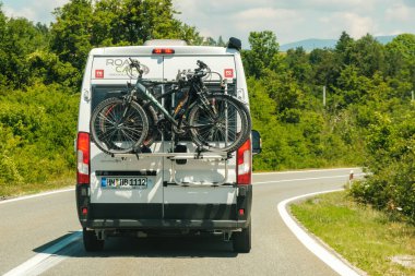 White passenger local bus drives on empty roadway past green trees on sunny day. White vehicle carries fastened sports bicycles on back closeup