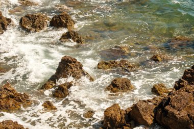 Small ocean waves crashing of rocks on shallow coastline at high tide at bright sunlight. Transparent waves create foams rolling on stones