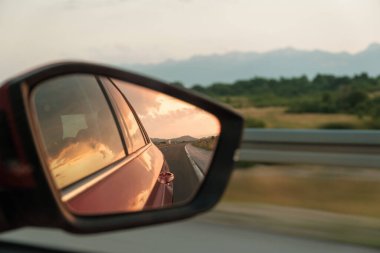 Red car drives past hilly landscape. Rear-view mirror shows roadway behind vehicle and door glass reflects sunset sky closeup