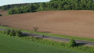 Motorbiker rides on empty road past yellow ripe field at sunset. Trees cast long shadows on road and field. Nice weather in countryside aerial view