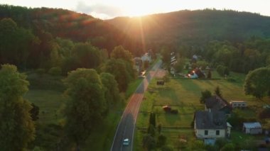 Old European village with small houses among forestry mountains. Long two-lane road stretches in rural area of highland valley at sunset aerial view